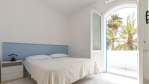 A bedroom with a bed, a nightstand, and a door leading to a balcony with palm trees.