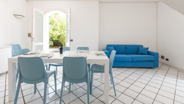 A modern living room with a white dining table and blue chairs, a blue sofa, and a large window with a view of greenery.