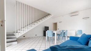 A modern living room with a blue sofa, a dining area with a white table and chairs, and a staircase with white railings.