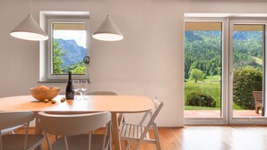 Uma sala de jantar com mesa de madeira, cadeiras brancas e vista para uma paisagem montanhosa através de grandes janelas.
