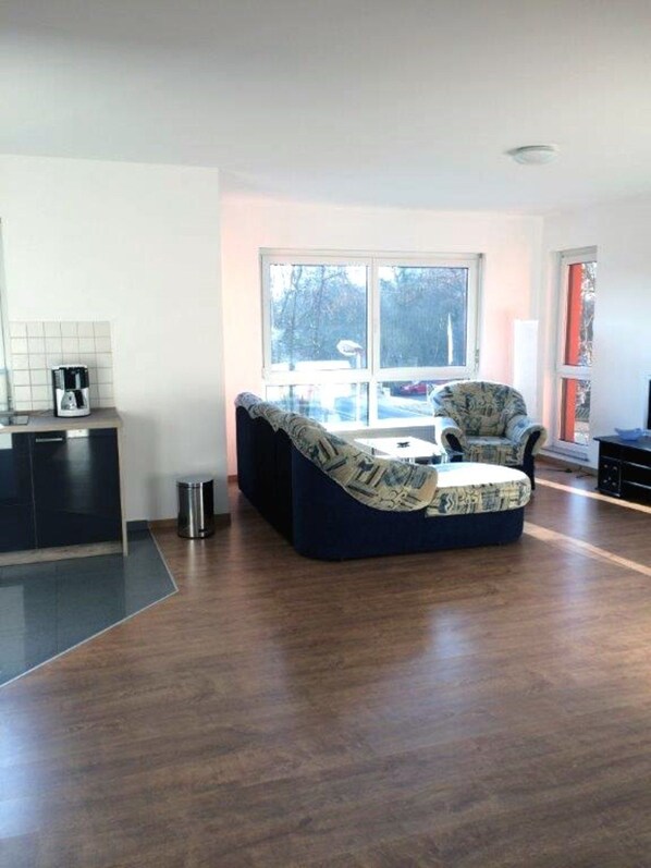 A modern living room with a large window, a sofa, a coffee table, and a kitchen area with a coffee maker.