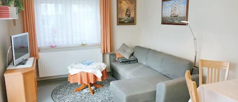 Uma sala de estar moderna com um sofá cinzento, uma mesinha redonda, uma TV em um armário de madeira e uma janela com cortinas.