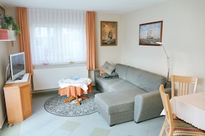 Ett modernt vardagsrum med en grå soffa, ett litet runt bord, en tv på ett träskåp och ett fönster med gardiner.