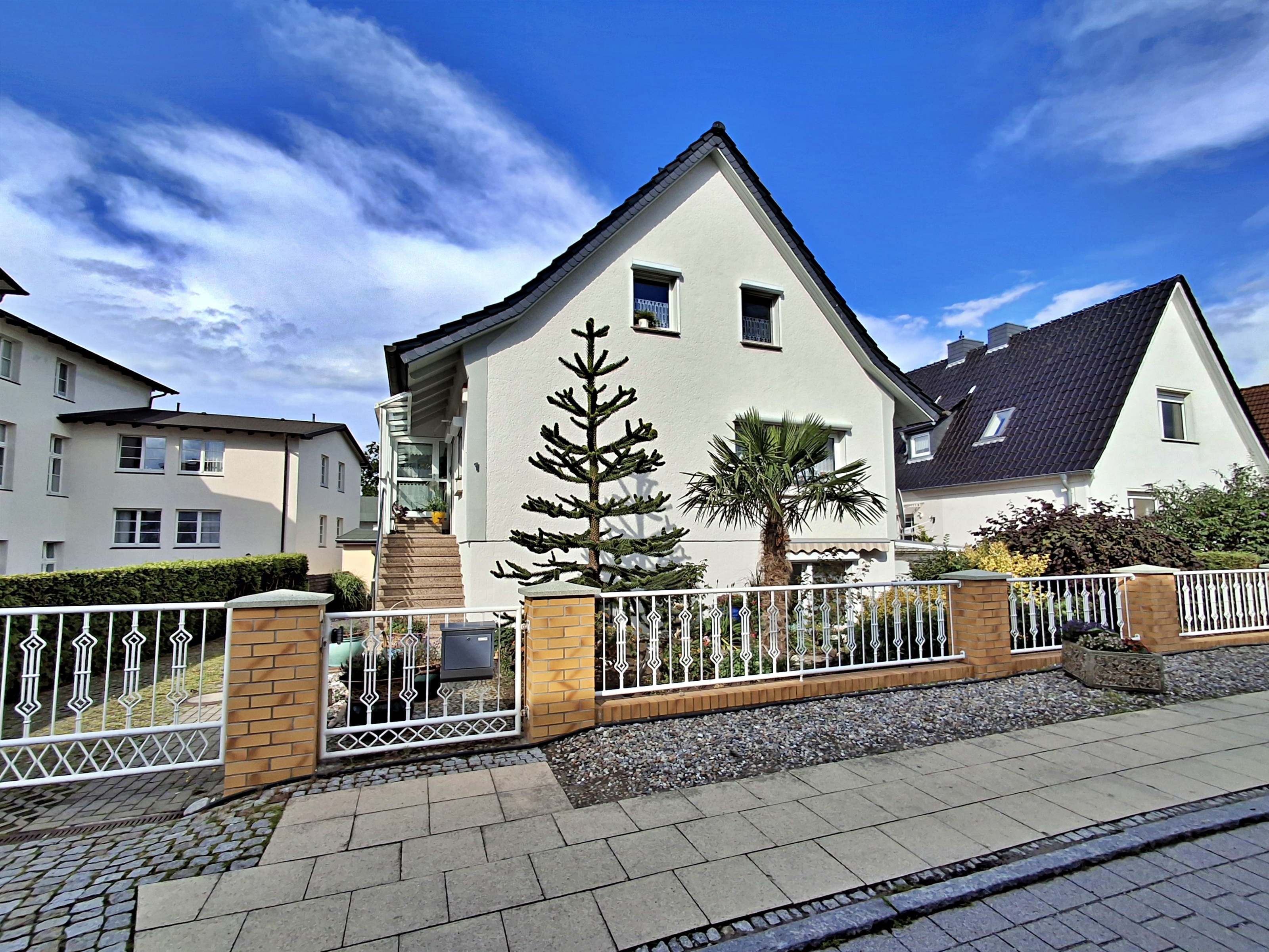 Une maison moderne à deux étages, avec une façade blanche, une entrée en briques et une clôture en métal.