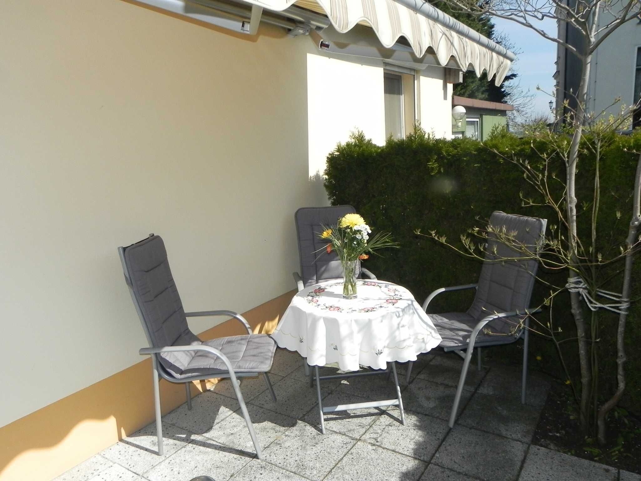 A patio with a table and two chairs, a vase with flowers, and a hedge in the background.