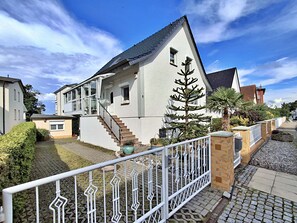 Une maison moderne à deux étages, avec une façade blanche, un escalier menant à l’étage supérieur et un petit jardin agrémenté d’un palmier et d’un cactus.