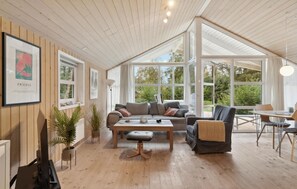 Relax in this inviting space with a comfortable sofa, dining table, and large windows offering views of the surrounding greenery.