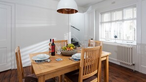 Uma área de jantar com mesa de madeira, cadeiras, uma tigela de frutas, uma garrafa de vinho e uma luminária pendente.