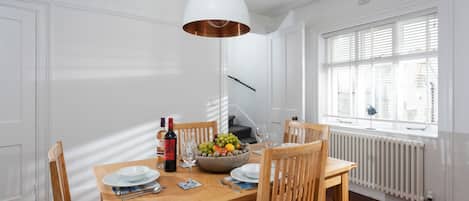 A dining area with a wooden table, chairs, a bowl of fruit, a wine bottle, and a pendant light.