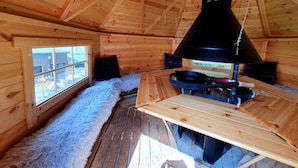 Interior de una cabaña de madera con chimenea, un banco cubierto de pieles y una ventana con vistas.