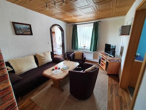 Uma sala de estar com sofá, poltronas, mesa de centro, televisão e uma janela com cortinas.