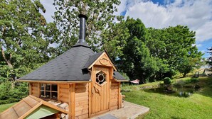Una cabaña de madera con chimenea, una pequeña terraza y un estanque al fondo.