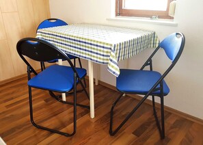 A room with a table and two blue chairs, wooden flooring, a window, and a plain wall.
