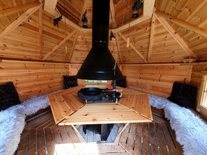 Interior de una cabaña de madera con una mesa central, una estufa negra y un ventanal.