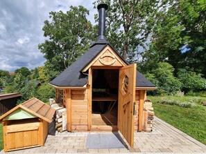 Una cabaña de madera con chimenea, una puerta abierta al interior y un cobertizo pequeño adyacente.