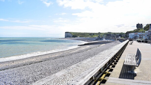 A pebble beach with a wooden walkway and benches, overlooking the sea and cliffs.