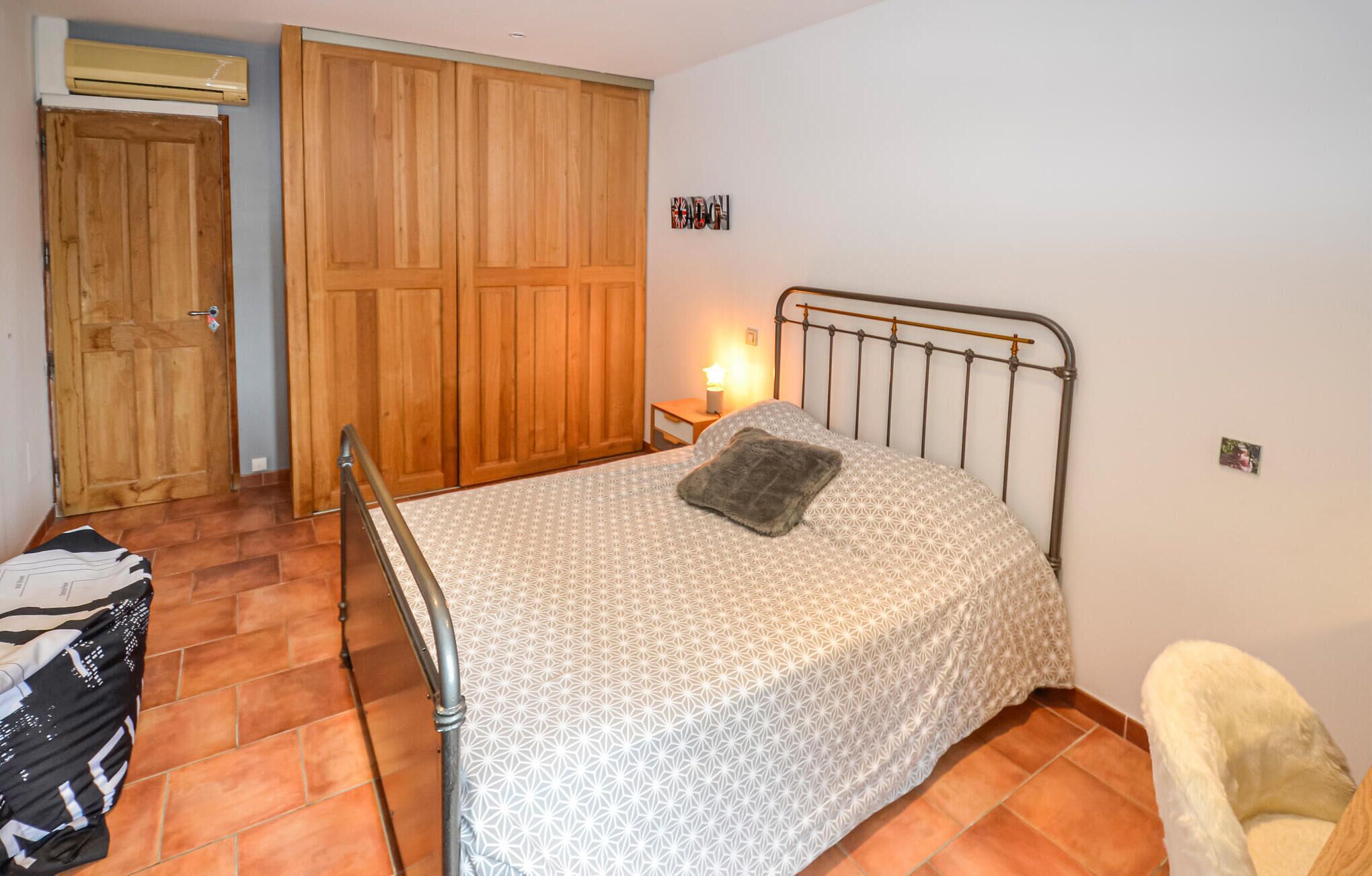 Ein Schlafzimmer mit einem Bett, einem Holzschrank, einem kleinen Nachttisch mit Lampe und einem Sessel.