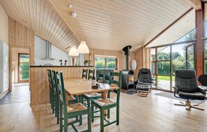 A spacious living area with a wooden ceiling, a large dining table surrounded by chairs, a fireplace, and large windows offering a view of the outdoors. - Beautiful home in Ansager with sauna (Ansager)