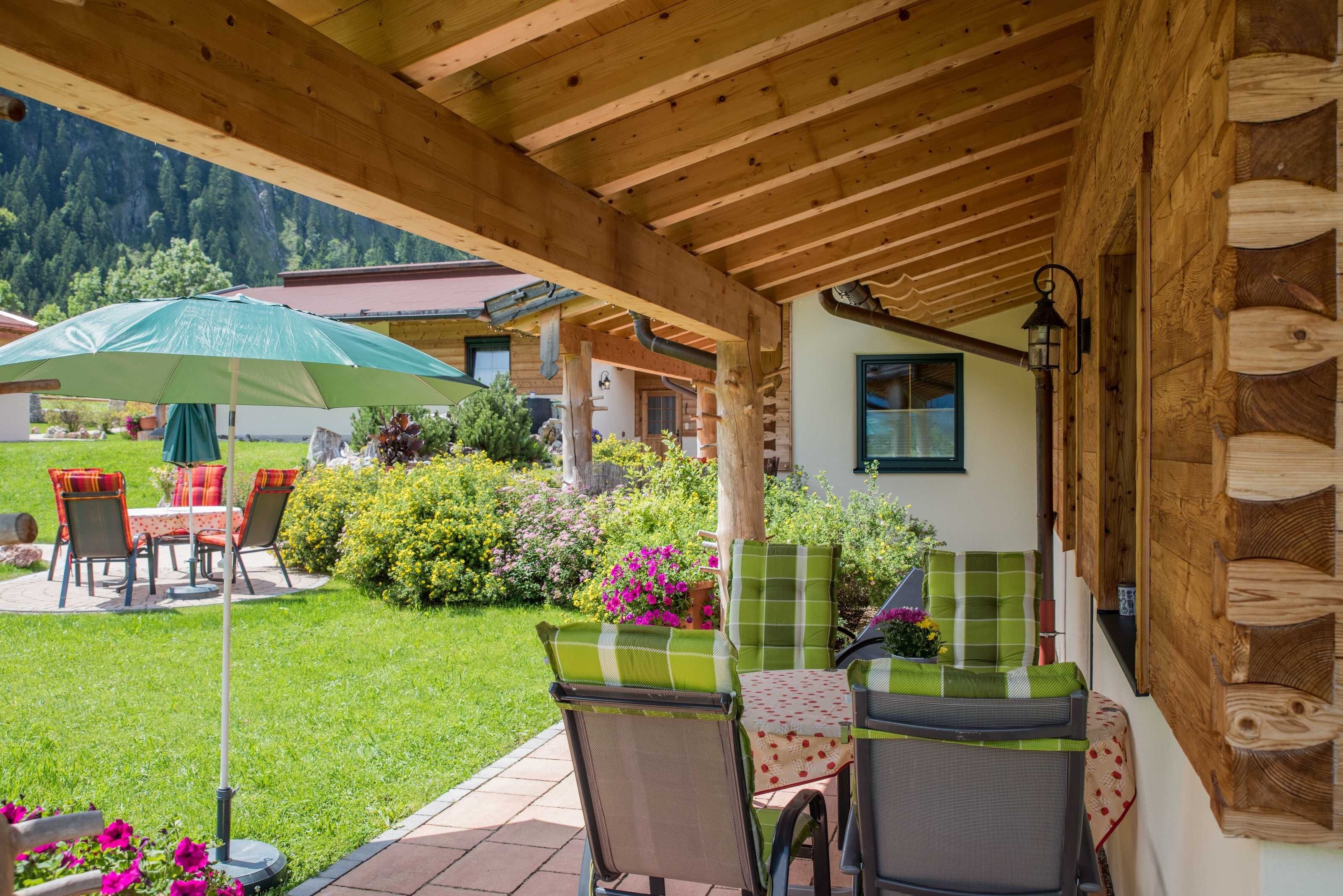 An outdoor patio with wooden beams, a table with chairs, a green umbrella, and a view of a mountain.