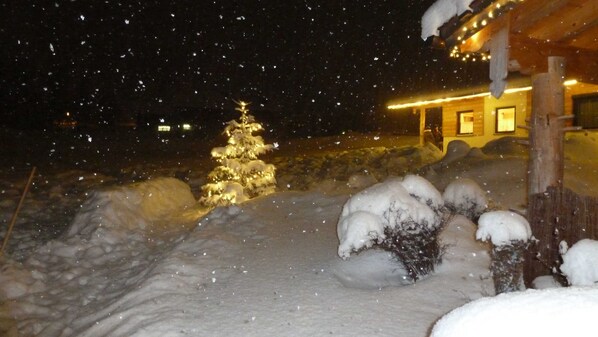 A snow-covered landscape with a lit Christmas tree and a building with a warm glow.