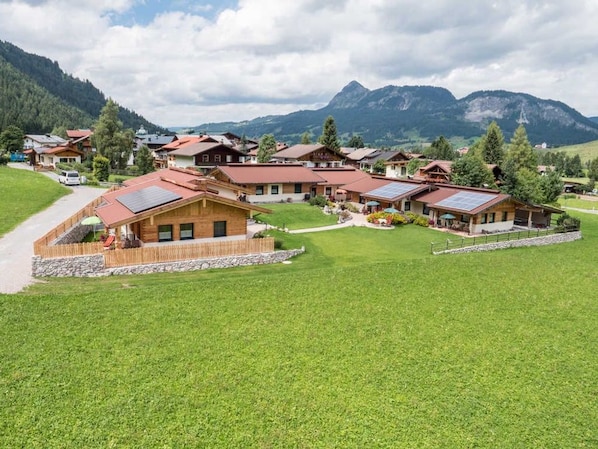 A cluster of wooden cabins with solar panels, surrounded by lush green fields and mountains in the background.