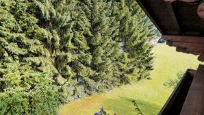 Vista desde un balcón de madera con vistas a un bosque frondoso y un área verde con algunas flores moradas.