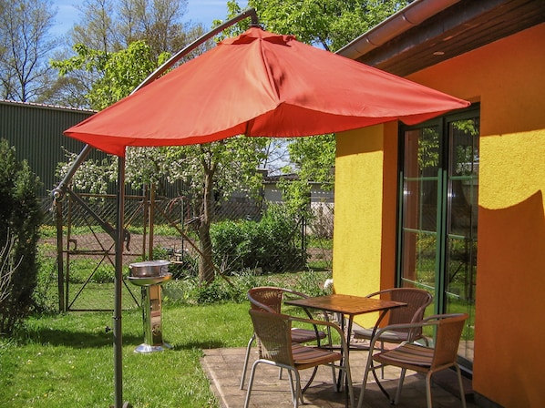 An outdoor patio with a table and chairs under a red umbrella, a metal grill, and a yellow wall with a window.
