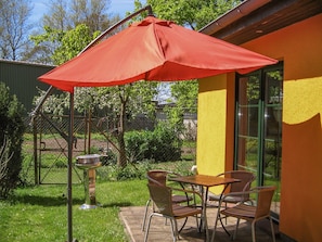 An outdoor patio with a table and chairs under a red umbrella, a metal grill, and a yellow wall with a window. - Vacation cottage Bohne, Germany (Kenz-Küstrow)