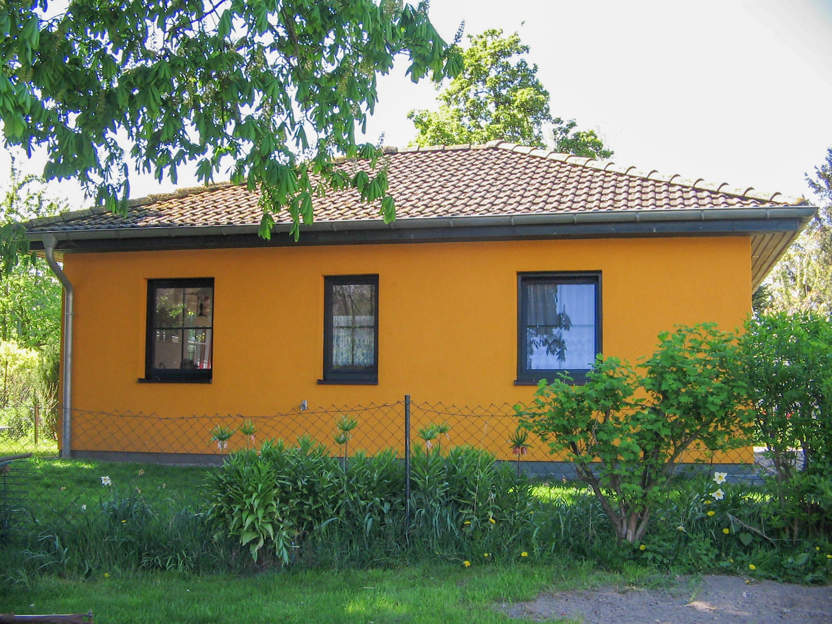Ein gelbes Gebäude mit einem Ziegeldach, drei Fenstern und einem Drahtzaun davor.