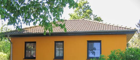 Un edificio amarillo con techo de tejas, tres ventanas y una cerca de malla metálica al frente.