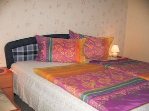 A bed with a floral-patterned bedspread and multiple pillows, a bedside lamp, and a nightstand with a lamp. - Vacation cottage Bohne, Germany (Kenz-Küstrow)