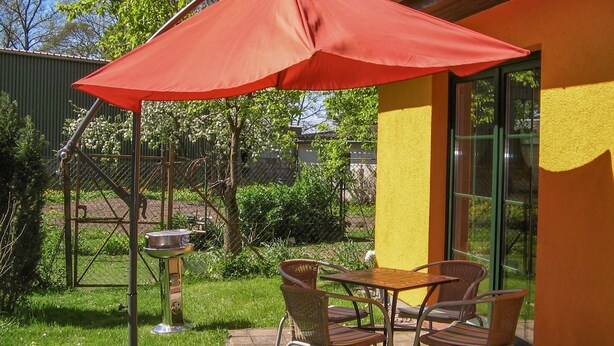 An outdoor patio with a table and chairs under a red umbrella, a metal grill, and a yellow wall with a window. - Vacation cottage Bohne, Germany (Kenz-Küstrow)