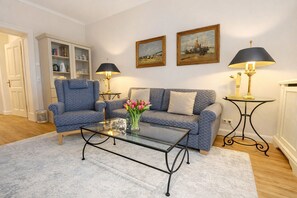 A living room with a patterned sofa, a glass coffee table with a vase of flowers, two lamps, and framed paintings on the wall.