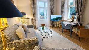 Un salon avec un canapé, une table basse et un téléviseur. Il y a des rideaux aux fenêtres et une lampe sur la table d’appoint.