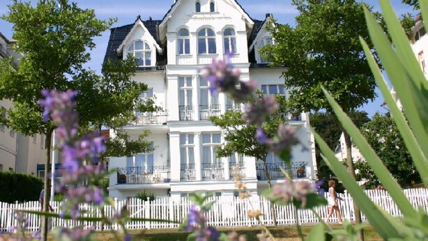 Un grand bâtiment blanc à plusieurs étages, doté d’une façade symétrique, entouré d’une clôture blanche en piquets et d’une végétation luxuriante.