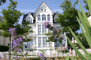 A large, white, multi-story building with a symmetrical facade, surrounded by a white picket fence and lush greenery.