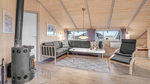A cozy cabin interior with a wooden ceiling, a sofa, a chair, a small table, and a wood stove. - Beautiful home in Ringkøbing with sauna (Ringkøbing)