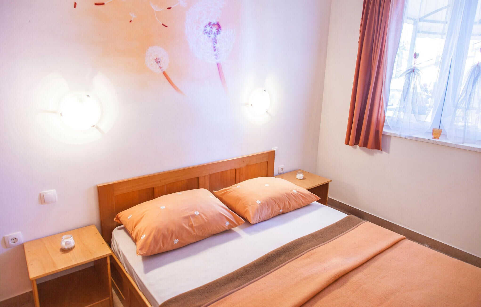 Un dormitorio con cabecera de madera, dos mesitas de noche, cama con ropa de cama naranja, una lámpara de pared y una ventana con cortinas.