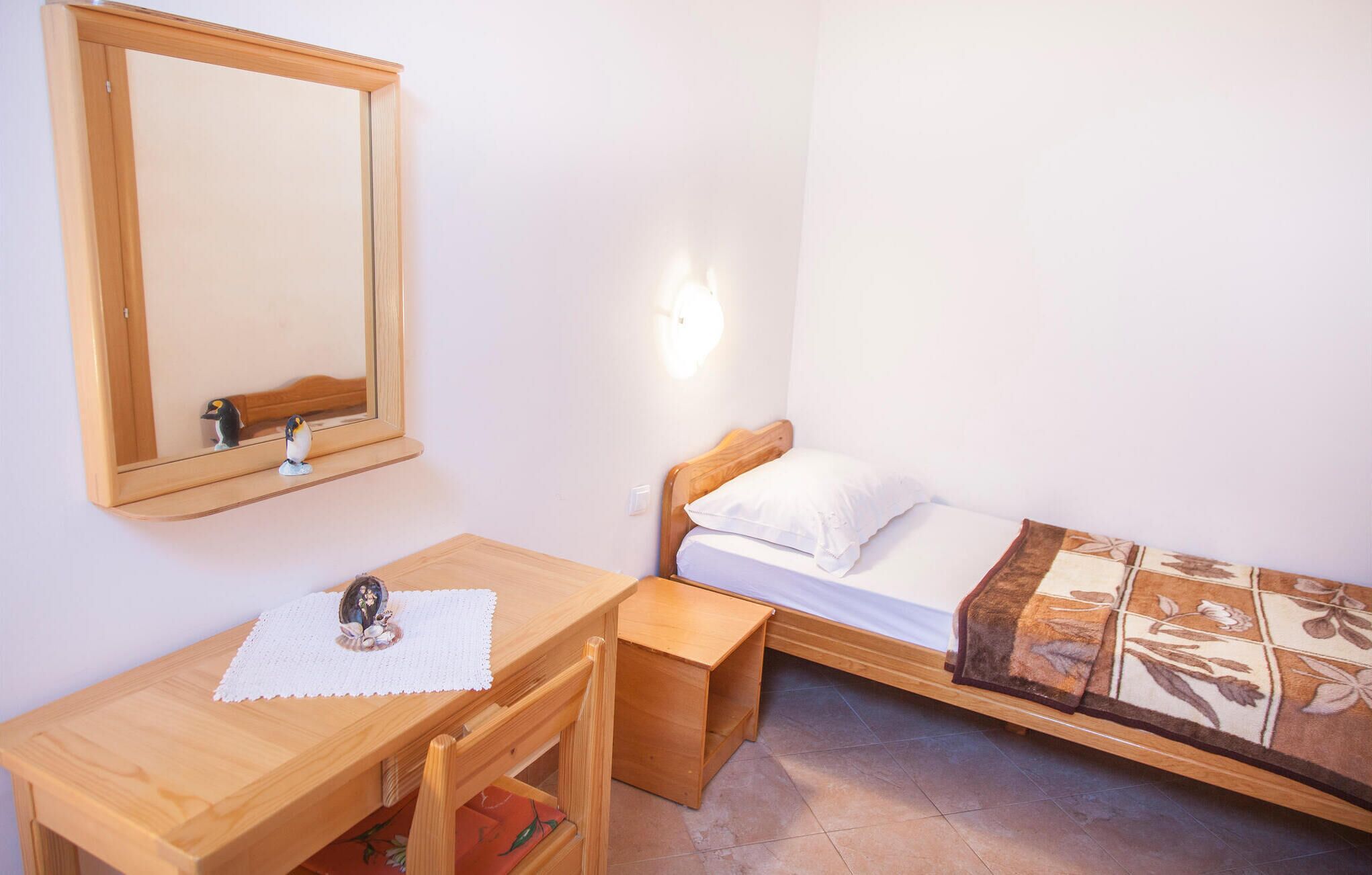 Quarto pequeno e arrumado, com escrivaninha de madeira, uma cama simples com manta estampada, mesa de cabeceira e espelho na parede.
