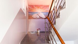 Una escalera con barandilla metálica y pasamanos de madera, que lleva a un rellano con un mural de un atardecer y conchas marinas.