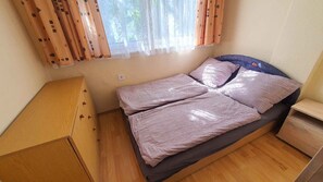 Una habitación pequeña con cama individual, piso de madera, una cama con colcha a rayas, una mesita de noche y cortinas.