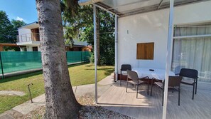 Un espacio de estar al aire libre cubierto con una mesa y sillas, un banco de madera, y vistas a una casa y una valla verde al fondo.