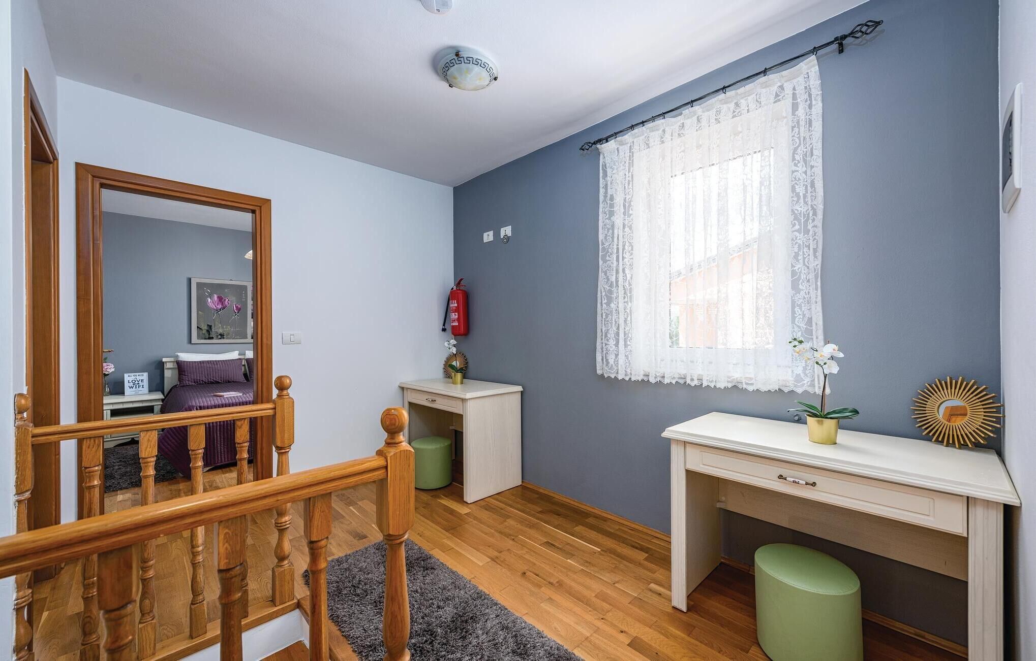 A bedroom with a wooden staircase, a bed with purple bedding, a bedside table with a plant, and a window with lace curtains.