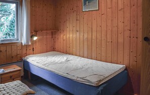 Ein kleines Zimmer mit einem Einzelbett, Holzwänden, einer Nachttischlampe, einem kleinen Tisch und einem Fenster mit Blick auf Bäume.