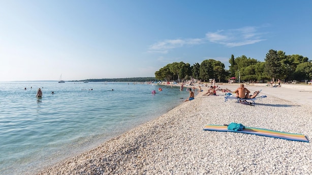 Une plage de galets avec une eau cristalline, des gens qui se baignent et prennent des bains de soleil, et un voilier à l’horizon.