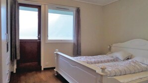 Ein kleines Zimmer mit einem Einzelbett, Holzboden, einem Fenster mit Vorhängen und einer Tür.