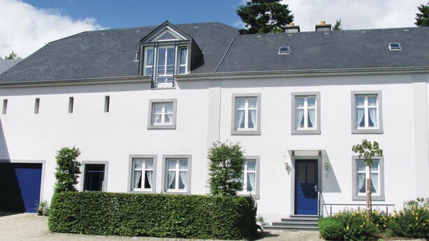 Ein weißes Gebäude mit grauem Dach, zahlreichen Fenstern und einer blauen Tür.