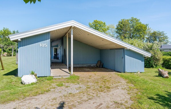 A small, single-story building with a sloped roof and a porch. It has a number "185" on the side and is surrounded by a grassy area with trees in the background.