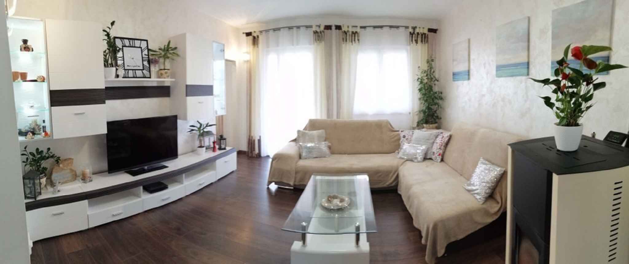 Een woonkamer met een televisie op een witte tv-kast, een beige bank, een glazen salontafel en een plant op een zwarte kast.