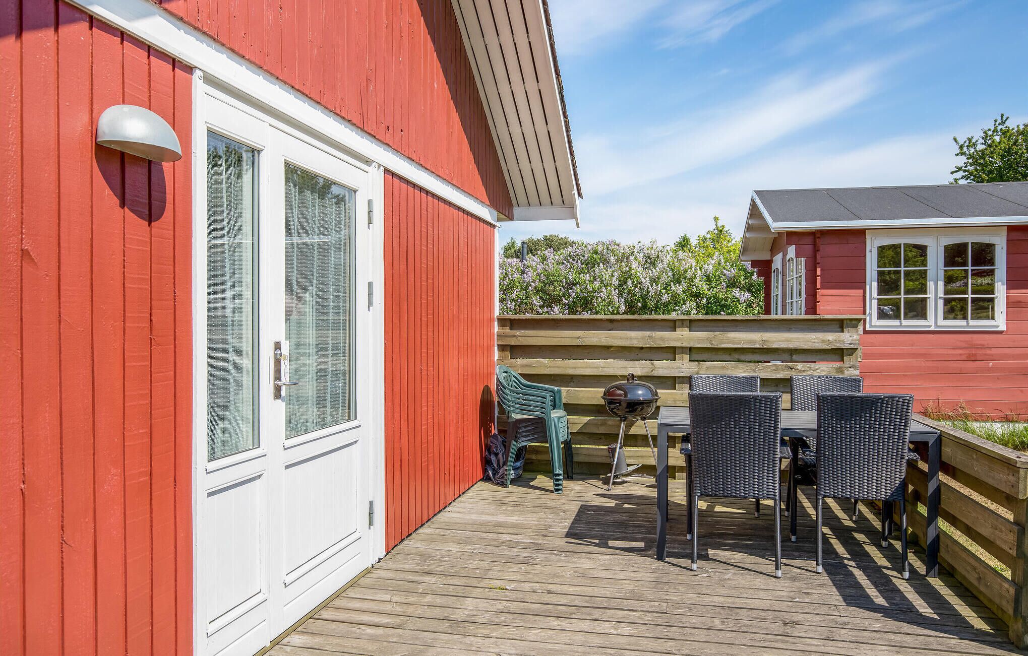 A red building with a white door and windows, a wooden deck with outdoor furniture, and a view of a garden with flowering plants.